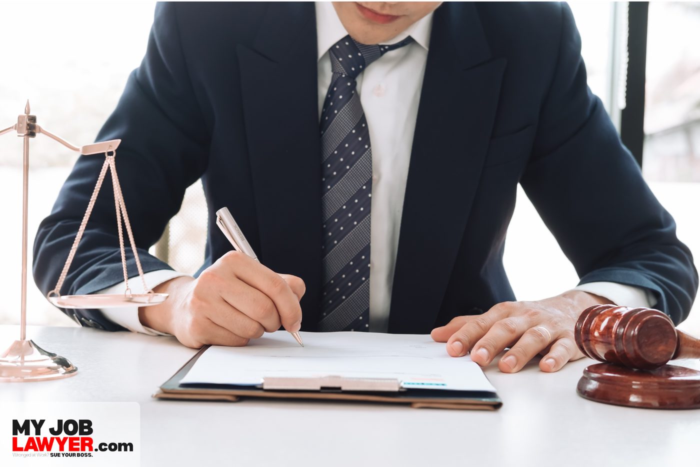 An attorney in a suit signs a document at his desk, next to a gavel and a set of scales of justice.