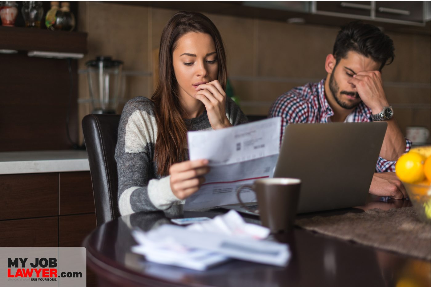 A worried woman reads a letter at the kitchen table while her partner holds his head in front of a laptop.
